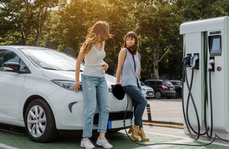 Two women lean on the hood of an electric vehicle as it charges.